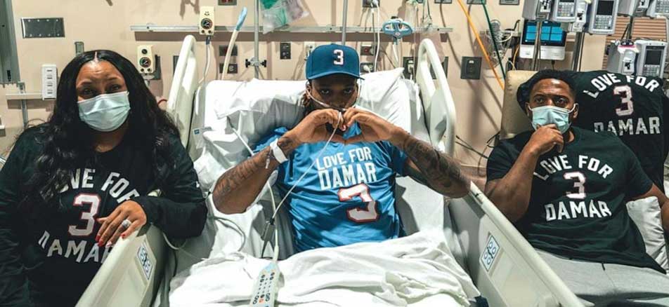 Damar Hamlin is hospital bed making heart sign with his hands. His mom is on his left and dad is on his right.